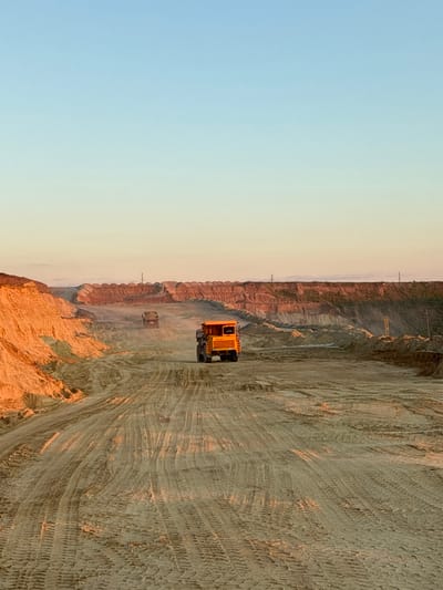 The image depicts an outdoor scene at a quarry or construction site, likely in Tatsinskaya, Russia, during what appears to be either sunrise or sunset, given the warm, golden light illuminating the sandy terrain and the clear, pale blue sky. The foreground is dominated by a wide, sandy track imprinted with tire tracks from heavy machinery. In the middle ground, two large, yellow dump trucks are visible. One is closer to the right side of the frame, and another, slightly smaller truck is further in the distance, positioned in the center of the track. The background features layered, sculpted mounds of earth and sand, characteristic of a quarry or excavation. Power lines and poles are intermittently visible along the upper edge of the distant mounds, suggesting proximity to infrastructure. The overall impression is one of an industrial or earthmoving operation in a vast, open environment, bathed in the soft glow of low-angled sunlight.