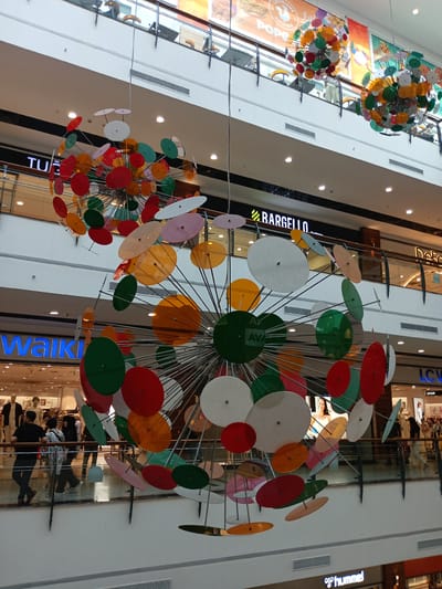 This is an eye-level, medium shot of the interior of a multi-level shopping mall. In the foreground, a large, spherical art installation constructed from metal rods and numerous colorful, flat, circular discs made of translucent plastic hangs from the ceiling. The discs are in shades of red, orange, green, pink, and white, and are arranged in a radial pattern. In the middle ground, people are visible on an upper level walkway, some standing and some walking. Several storefronts are visible behind them, with illuminated signs such as "TUĞBA", "BARGELLO", "WALKWAY", "LCV COÇUK", and "hummel". The mall appears to be modern, with white walls and railings. The context provided indicates the location is the NeoPlus Outlet mall in Kırıkkale, Turkey.