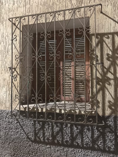 This is a medium shot, eye-level, outdoor photograph of a window with decorative wrought iron bars and shutters, likely in the city of Berrechid, Morocco. The window is set into a textured, beige stucco wall. Below the window, there is a rough, grey textured surface. The wrought iron bars are arranged vertically and diagonally, with decorative scrollwork at the intersections. The shutters behind the bars are closed and appear to be made of dark wood with horizontal slats. Sunlight casts shadows of the wrought iron bars and possibly foliage onto the wall and the ground below. A thin green string is tied to one of the bars. The overall scene suggests a residential or commercial building in an urban environment. There are no people or animals visible in the frame.
