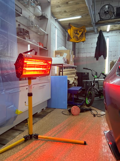 The image depicts an interior scene of what appears to be a workshop or garage in Veteran-2, Russia. The room has white cinder block walls and a wooden plank ceiling. In the foreground, a bright orange infrared heat lamp on a yellow stand is actively emitting heat, casting a warm red glow on the floor. To the left, a large blue plastic sheeting is draped, possibly for protection or containment.

Shelving units filled with various items, including bottles and containers, are visible against the wall. A fan is mounted on the ceiling. Various tools and equipment are scattered around the space, including a bicycle with green accents, a polishing machine with a pad, and stacks of firewood. A portion of a car is visible on the right side of the frame, its metallic surface reflecting the ambient light. The overall impression is one of a functional workspace with an ongoing task, likely related to automotive detailing or repair, indicated by the presence of the heat lamp and polishing equipment. There are no people visible in the scene.