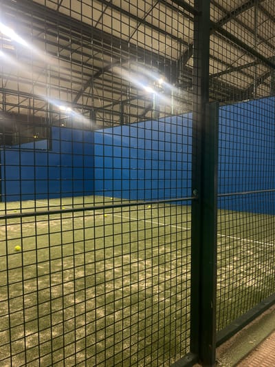 The image shows a view of a padel tennis court from behind a metal fence. The court is split into two halves, one with a blue surface and the other with a green surface. Several tennis balls are visible on the green side. A building with a metal structure is seen through the fence, reflecting sunlight. The location is the city of Alcobendas, Spain.