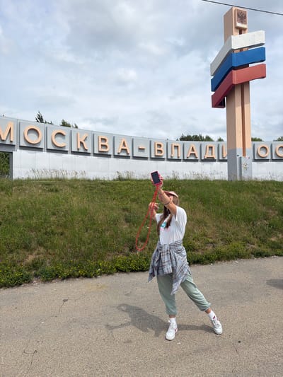 The image shows a young woman taking a selfie in front of a large sign that reads "МОСКВА-ВЛАДИОСТОК" (Moscow-Vladivostok). The sign is composed of large, light-colored letters with a copper outline, mounted on a grey panel. To the right of the sign is a tall, beige pylon with rectangular blocks in the colors of the Russian flag (white, blue, and red) stacked horizontally, and a crest at the top. The woman is wearing a pink hat, a white t-shirt with a graphic print, a plaid shirt tied around her waist, light green sweatpants, white socks, and white sneakers with pink accents. She is holding a pink smartphone in her right hand, with a red lanyard attached. She is standing on an asphalt surface, with a grassy embankment with small yellow flowers behind the sign. The sky is overcast. The location is the city of Nikolaevka, Russia, at the sign marking the beginning of the highway to Vladivostok.