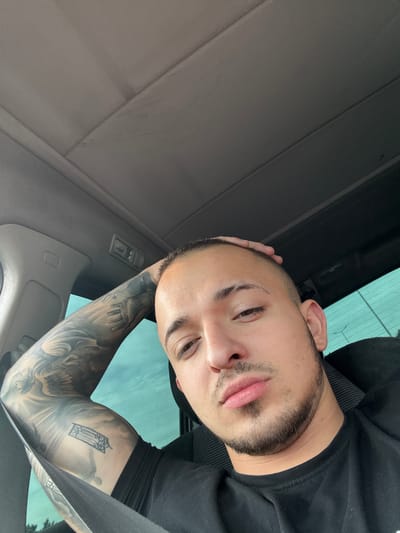 A man with a short buzz cut and light stubble on his chin is seated in a car, resting his left arm on the headrest and his hand on his head. He is wearing a black t-shirt and has extensive tattoos on his left arm, including what appears to be a religious or angelic figure and a smaller, more geometric tattoo. The interior of the car is visible, with the grey headliner and a window with a greenish tint. The lighting suggests it is daytime. The image is a selfie taken from a low angle. There is no information about the location in the city of Stroevo, Bulgaria, in the image.