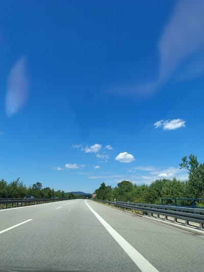 The image is a highway scene captured from the perspective of a vehicle in motion on a clear, sunny day. The sky is a vibrant blue with scattered white clouds. The highway is paved and marked with white lane lines. A metal guardrail runs along the right side of the road. Dense green trees and foliage line the sides of the highway. In the distance, a gentle hill with trees is visible. There are several cars on the highway, all appearing to be driving in the same direction as the viewpoint. The scene is devoid of people. The image is taken in Niederwinkling, Germany.