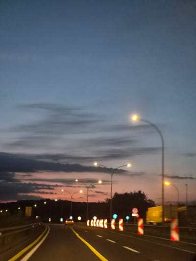 The image depicts a highway in Mondercange, Luxembourg, at dusk. The sky is a gradient of deep blue to pale blue, with streaks of dark clouds tinged with pink from the setting sun. A line of streetlights illuminates the road, casting a warm glow. The road itself has bright yellow dividing lines. Several cars are visible on the highway, including a red car in the middle distance and a yellow truck to the right. Construction barriers, marked with red and white reflective stripes, are present along the side of the road, suggesting ongoing work. Trees and vegetation form a dark silhouette against the horizon. The scene is quiet and tranquil, characteristic of a highway at the end of the day.