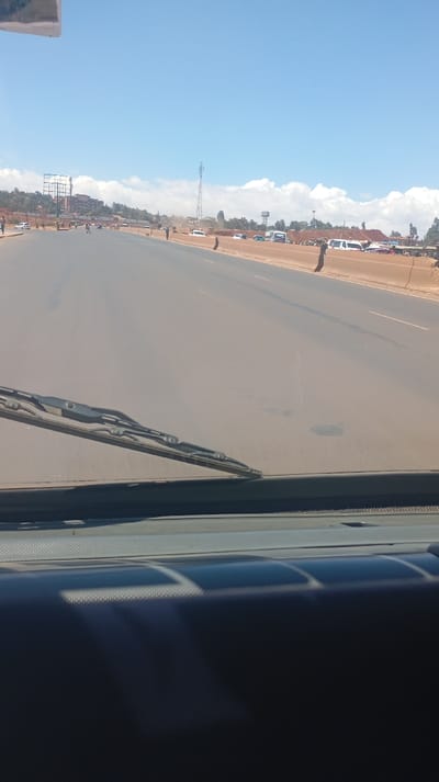 A view from inside a vehicle driving on a multi-lane road. A windshield wiper is visible in the foreground. The road is dusty and dry, with construction barriers on the side. In the distance, there is a billboard, buildings, and a cell tower. A line of vehicles is visible on the side of the road. The sky is bright blue with some clouds.