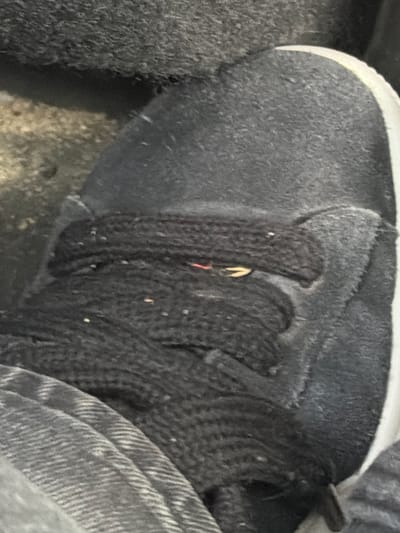 The image shows a close-up view of the top of a dark-colored shoe with black laces. The shoe appears to be made of suede and has a white accent line along the edge. The laces are thick and knotted. There are some small white specks on the laces and the shoe, possibly dust or debris. A small red and yellow object is visible between the laces, possibly a piece of string or a small foreign object. The background is dark and textured, suggesting an outdoor or industrial setting. The shoe is likely located on a street or pathway in Vallensbæk Landsby, Denmark.
