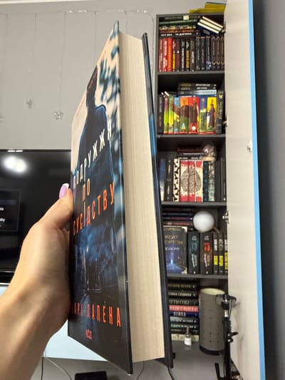 A hand with a manicured nail, painted with white polish and a hint of purple, is holding a thick book with a dark blue cover featuring an orange title. The book appears to be titled "Подружки по сусідству" or a similar Cyrillic script. The background shows a bookshelf filled with many books. One shelf contains colorful spines, while others are filled with books of various colors and sizes. A small, spherical white object is visible on one shelf, and a partially opened cabinet door is on the right side of the image. The environment suggests an indoor setting, possibly a room with a TV or monitor visible in the blurred background to the left. There is no indication of the city of Łódź, Poland.