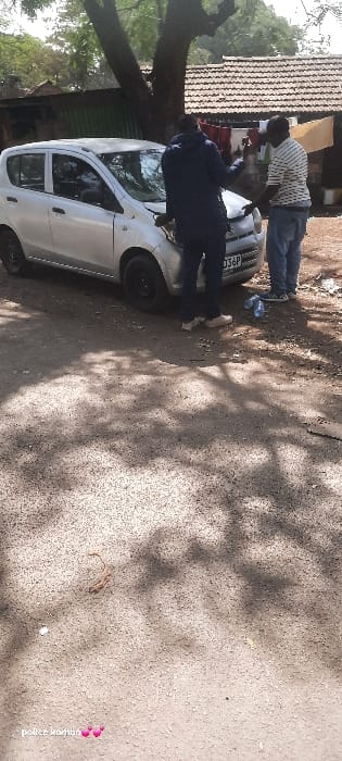 In the city of Homa Bay, Kenya, an outdoor scene depicts two adult males and a silver five-door hatchback vehicle on a light-colored, unpaved surface. The vehicle, bearing Kenyan license plate KDG 036P, is positioned with its front facing the viewer's right, exhibiting minor front-end damage or a slightly misaligned hood on the passenger side.
The first male, on the left, wears a dark blue long-sleeved top and light-colored trousers/shoes, facing the second male with his left hand raised in a gesture. The second male, on the right, wears a white and light-striped short-sleeved polo shirt, blue jeans, and dark athletic shoes, facing the first male with his right hand extended towards the vehicle's front. Both individuals appear to be engaged in conversation or inspection of the car's front section.
Scattered debris, including a clear plastic bottle, is visible on the ground near the vehicle. The background features a large tree, a dark green corrugated fence to the left, and several single-story buildings with reddish-brown tiled roofs. Laundry is observed hanging on a line against one of the buildings. The scene is illuminated by strong sunlight, casting distinct shadows from the tree and individuals onto the ground.