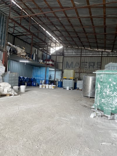 An interior view of a large, open warehouse space in the village of Kenen Azirbayev, Kazakhstan, is captured. The structure features a corrugated metal roof supported by exposed steel beams, with natural light filtering through gaps in the roofing. The floor is a rough, concrete surface, bearing marks and scuffs indicative of heavy use.
The warehouse is filled with various industrial containers and materials. To the left, large, white plastic IBC totes (intermediate bulk containers) are stacked, and further to the left, several large white bags of material rest on a pallet. In the mid-ground, a row of blue-lidded drums and smaller buckets are lined up. To the right of these, several white buckets, some appearing to be paint cans, are arranged. Behind them, more drums, some red, are visible on a raised shelf.
Dominating the right side of the image are several large, cylindrical industrial containers. A prominent green plastic tank, heavily splattered with white material, is in the foreground. Behind it, a large stainless steel cylindrical tank is visible, and further back, more IBC totes, some clear and some light yellow, are stacked. The back wall of the warehouse appears to be constructed from corrugated metal shipping containers, with large, faded letters spelling "MERS" visible on the surface.
No people are present in the scene. The overall impression is one of an active, though perhaps somewhat cluttered, industrial storage or processing facility.