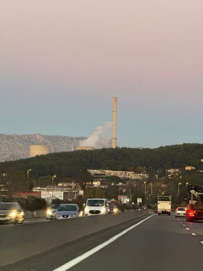 A tall chimney rises into the sky near a mountain range. White smoke billows from the chimney, partly obscuring the mountain. A highway with several vehicles runs parallel to a forest. Streetlights illuminate the scene, with advertisements and signs alongside the road. The sky is a gradient of colors, transitioning from a pinkish hue to a pale blue. The entire image is oriented sideways.