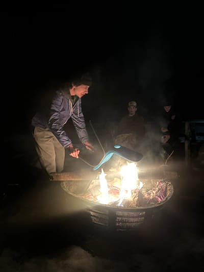 A man in a blue jacket and beige pants is outside at night. He holds a blue chair in his hands, seemingly about to throw it into a large fire. The fire is contained within a metal ring, possibly a grill or fire pit, filled with burning debris. There is another person standing behind the man, facing the fire. A string of objects dangles above, near a dark structure. The scene is dark, with smoke rising.