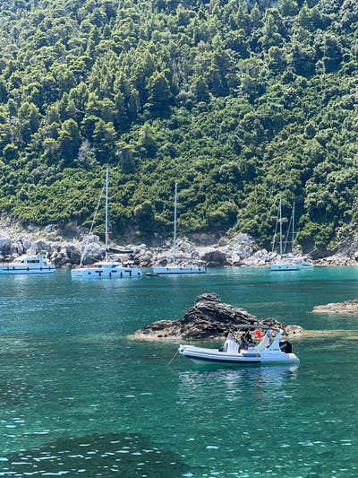 The image depicts a serene coastal scene in Loutraki, Greece. The foreground features crystal-clear turquoise water, with sunlight creating dappled patterns on the seabed. A white inflatable boat with a black canopy is anchored in the mid-ground, carrying two individuals, one of whom is steering. The boat is labeled "AL MARAH". In the background, several white sailboats are anchored, their masts reaching towards a lush, green hillside densely covered with trees. Rocky outcrops punctuate the shoreline. The overall impression is one of tranquility and natural beauty.