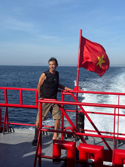 A middle-aged Vietnamese man, wearing a black sleeveless shirt and khaki shorts, stands on the deck of a red-railed boat, looking to his left with a slight smile. A Vietnamese flag is prominently displayed, waving in the breeze. The boat is moving, evidenced by the wake trailing behind it in the deep blue ocean. The sky is clear and blue, with a hint of distant land on the horizon. The scene appears to be a sunny day at sea, possibly near the coast of Dong Son, Vietnam.