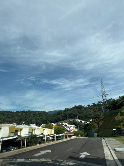 This image depicts a residential area within Nuevo Cuscatlán, El Salvador, characterized by a sloping asphalt road leading uphill, bordered by concrete curbs. In the immediate foreground, white painted arrows on the road indicate a straight direction and a left turn. The bottom edge of the frame shows a reflection, likely from a vehicle's dashboard.
On the left side of the road, a series of modern, multi-story houses are visible. These structures are predominantly white with yellow-accented sections and feature open carports beneath them, accommodating several parked vehicles. One carport exhibits an installed solar panel array.
To the right of the road, a steep, terraced hillside covered in green grass ascends, with visible dark green retaining structures or barriers. A prominent metal lattice utility tower, supporting power lines that traverse the upper mid-ground, stands on this hillside.
The mid-ground features further residential development with white and light-colored houses nestled into the surrounding hilly, densely forested terrain. The background is dominated by a range of tree-covered mountains or hills. The sky is partly cloudy, displaying a mix of blue sky and scattered white to light grey cloud formations. No individuals are visible.