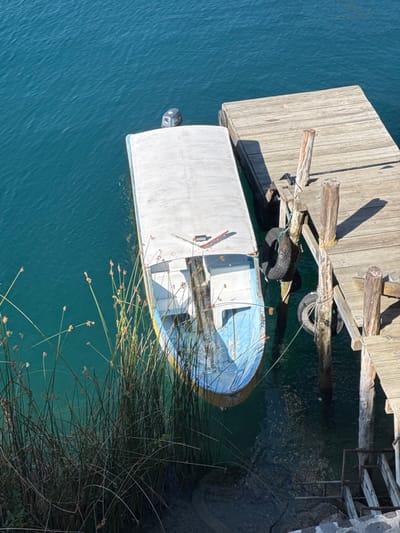 The image presents an aerial-level view of a boat moored at a wooden pier on a body of water in San Pablo La Laguna, Guatemala. The water, likely Lake Atitlán, is a clear, teal-blue color, occupying the left and upper sections of the frame, exhibiting minor surface ripples. In the foreground, along the left edge, a dense cluster of tall, thin green reeds with small, brownish seed heads rises from the water, partially obscuring the immediate shoreline.

A light-colored wooden pier, constructed of aged planks, extends from the right side of the frame into the water. It is supported by multiple weathered vertical wooden posts, some exhibiting light-colored rope tied around them. Two dark, worn rubber tires are suspended as bumpers from the pier posts. Below the pier on the lower right, a series of stone steps with metal railings descends into the water, suggesting an access point.

A single, elongated motorboat is moored parallel to the pier. The boat features a light-blue hull with a yellow stripe visible along its side, and a white, flat canopy covers its main passenger area. The bow is visible, showing an open interior with seating. An outboard motor is partially submerged at the stern of the boat. Ropes secure the boat to the pier posts. The scene is illuminated by bright daylight, evidenced by distinct shadows cast by the pier posts and the pier itself onto the water and wooden surfaces. No individuals are present in the photograph.