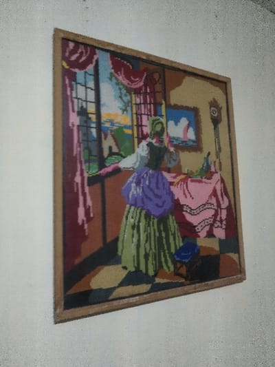 The image displays a framed textile artwork, likely a needlepoint or tapestry, mounted on a light off-white or light gray textured wall. The artwork depicts an interior scene with a woman standing by a window, gazing outdoors.

**Artwork Content:**
*   **Central Figure:** A woman is shown from the back and slightly to her left, wearing a white blouse, a dark green bodice, a voluminous purple overskirt, and a pleated green underskirt. Her hair is covered by a green headpiece. She leans on a dark ledge or windowsill, looking out the window to the left.
*   **Window View:** The window reveals an exterior landscape featuring a white lighthouse, various colorful buildings with red roofs, a body of water (possibly sea or lake), and a sky with hues of blue, orange, and yellow, suggesting a sunrise or sunset.
*   **Interior Details (Left):** Red and pink curtains with ruffled edges frame the window.
*   **Interior Details (Right):**
    *   A table is positioned to the right of the woman, covered with a pink tablecloth featuring white trim. On the table are several pieces of fruit (appearing as apples or oranges) and a dark bottle or pitcher.
    *   Above the table, a smaller framed picture hangs on the wall, depicting a sailboat on water under a blue sky with white clouds.
    *   Further to the right, a tall, dark grandfather clock with a visible pendulum is against the wall.
    *   Near the woman's feet, a small blue stool or footrest is present.
    *   The floor features a checkered pattern of dark brown/black and tan squares.
*   **Overall Artistic Style:** The artwork utilizes a pixelated or stitched aesthetic characteristic of needlepoint, with bold, distinct color blocks.

**Physical Presentation:**
*   The artwork is enclosed in a simple, light brown wooden frame.
*   The surrounding wall is uniform in color, showing minor imperfections and a subtle texture, consistent with a painted interior wall.
*   The photograph is taken at a slight angle, with the right side of the frame appearing closer to the viewer.

**Location:**
The described framed artwork is presented as being located in the city of Chimay, Belgium. The image itself does not provide visual cues to confirm this specific geographic location, nor does the artwork depict scenes identifiable as Chimay.