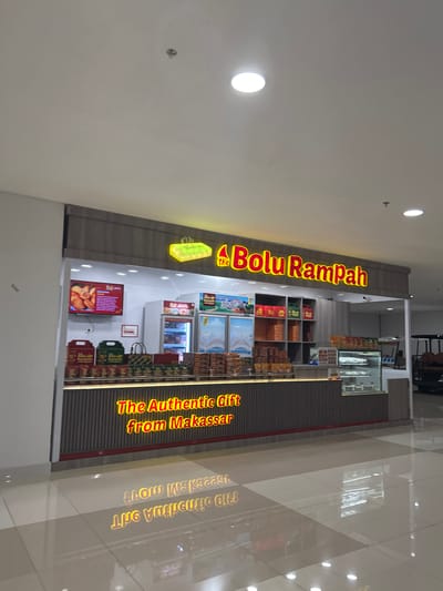 The image shows a brightly lit storefront for "Bolu Rampah" in the city of Baji Mangngai, Indonesia. The store's name is displayed prominently in large, illuminated red lettering. The storefront features a counter with display cases showcasing baked goods and shelves stocked with packaged products. A television screen on the left displays food items. The flooring is a light-colored tile with a polished finish, reflecting the lights from the ceiling. The overall environment appears to be an indoor shopping area.