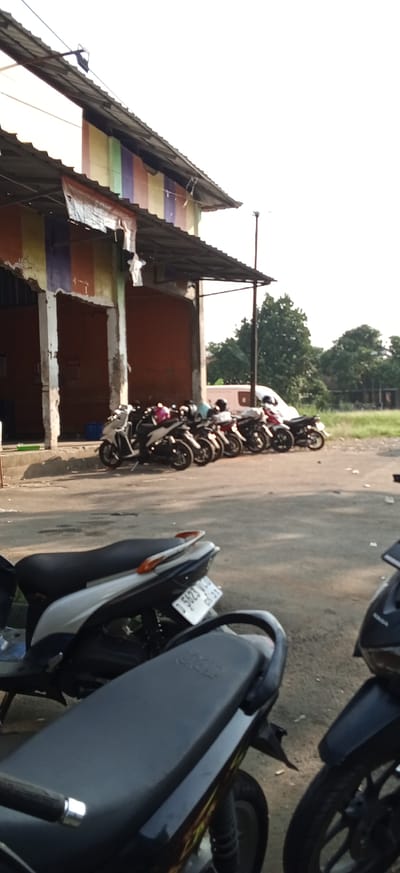 The image depicts a street scene in Kiangroke, Indonesia. In the foreground, the seats of two motorcycles are visible, one black and one black and silver. The black and silver motorcycle has a white license plate with the characters "D 5623 DUC". In the background, a row of parked motorcycles is lined up in front of a building. The building has a corrugated metal roof with colorful fabric panels hanging from the underside. The walls of the building are painted orange. Behind the building, trees and a bright sky are visible. A white van is parked to the right of the motorcycles. The overall impression is one of a street or market area, likely during the daytime.