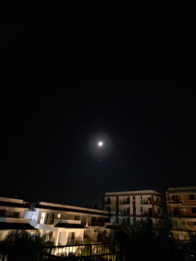 This image depicts a nighttime scene in Trachoni, Cyprus. The dark sky occupies the upper portion of the frame, featuring a prominent, bright moon centrally located, with a smaller, distinct celestial body positioned directly below it and another faint point of light to the upper left.
In the lower half of the frame, a series of multi-story buildings are visible, illuminated by artificial light sources from within and ambient moonlight. To the left, a light-colored, multi-story building complex with dark, sloping roofs exhibits several illuminated windows, indicating internal lights. An external metal staircase is visible on this structure. In the central area, a modern, light-facade multi-story building with multiple balconies features several windows glowing with a warm, orange internal light. To the right, a darker, multi-story structure appears partially constructed or in a state of disrepair, with visible exposed brickwork or concrete and an uneven facade. The immediate foreground shows the dark silhouettes of palm-like trees and the top railing of a fence or balcony. No individuals are visible, and no specific actions or interactions are occurring within the scene.