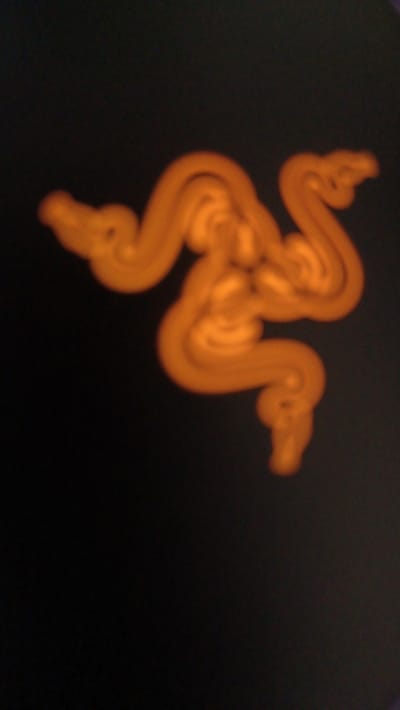 The image shows a blurry, bright orange Razer logo on a dark, unidentifiable surface. The location is a dark room, likely indoors, in Magelang, Indonesia, at the coordinates -7.479, 110.222. No people or other objects are visible in the image. The Razer logo is illuminated and stands out against the dark background. The image appears to be taken with a mobile device, likely at a low angle.
