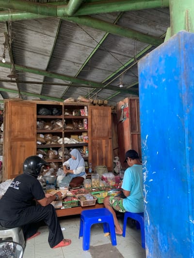 The scene is set inside a small, open-air market in Kecamatan Piyungan, Indonesia (latitude: -7.837, longitude: 110.471). The roof is constructed of corrugated metal, and the walls are composed of wooden panels and a large, blue, metal wall. A large wooden cabinet with several shelves is filled with bags of various spices and herbs. There are three people present. A woman in a white headscarf is seated on the floor, sorting and arranging ingredients in front of the cabinet. A man wearing a black t-shirt and a black helmet is seated on a stool to the left of the woman. He is interacting with items on a table in front of him. To the right of the woman is a man seated on a stool, wearing a turquoise shirt and camouflage shorts. He is interacting with items on the table in front of him. There are two blue plastic stools and a variety of small, plastic containers filled with different types of food items on the tables in front of the three individuals.