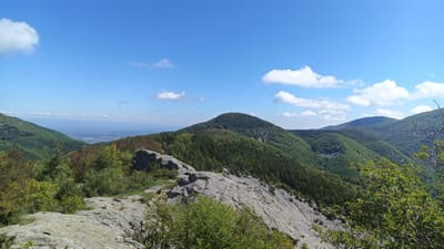 The image is a landscape photograph taken from an elevated rocky terrain, showcasing the mountainous region of Sini Vrah, Bulgaria. The foreground features gray rock formations and sparse vegetation. The midground shows rolling hills densely covered with green forests. In the background, there is a clear blue sky with scattered white clouds. A distant valley is visible between the mountain ranges.
