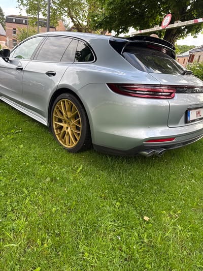 A silver Porsche Panamera is parked on green grass in an outdoor setting. The car features gold-colored rims. The license plate shows "1-HML-361". The location appears to be Melreux, Belgium, as evidenced by the license plate. A red and white sign is attached to a white metal barrier in the background. Buildings and trees are visible in the background. The weather appears to be overcast.