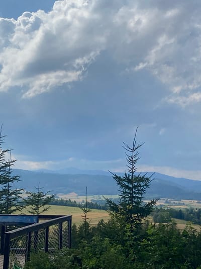 The image depicts a scenic vista of rolling hills and mountains under a cloudy sky. In the foreground, there is a dark wooden deck or platform with a railing made of dark metal poles and wire mesh. Several evergreen trees are visible, with a prominent one in the mid-right foreground. Beyond the platform and trees, a landscape unfolds with fields, some appearing to be golden or light brown, suggesting they might be harvested or dry grass. In the distance, layers of blue-toned mountains recede into the horizon. The sky is a mix of light blue and large, white, and gray clouds, some of which are dense and dramatic. The overall impression is one of a natural, rural landscape. The provided metadata indicates the location is Chełmsko Śląskie, Poland. There are no people visible in the image.