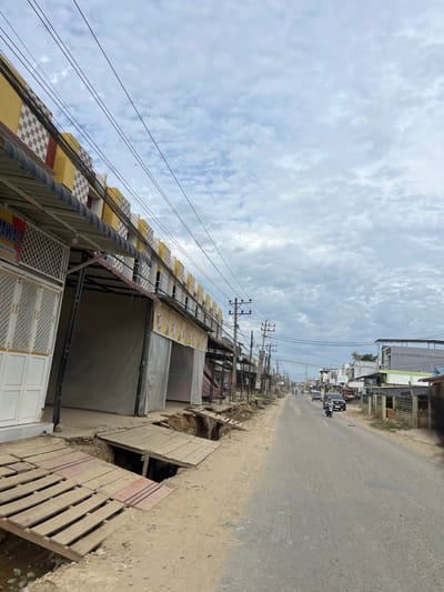 The image depicts a street scene in Landuh, Indonesia, characterized by overcast skies and a slightly dilapidated urban environment. The foreground shows a section of a building with a yellow and white checkered facade and a corrugated metal awning. In front of this building, the ground has eroded, creating a large sinkhole. Temporary wooden plank bridges have been constructed across the damaged area to allow pedestrian access.
The street itself is paved with asphalt, showing signs of wear and tear with visible potholes. On the left side of the street, a row of buildings with awnings and closed storefronts lines the road. Overhead, a network of electrical wires and utility poles crisscross the sky, typical of many Southeast Asian urban areas.
Further down the street, other buildings are visible, some with signage, including one that appears to be a "BSI" bank. Traffic consists of a few vehicles, including cars and motorcycles, moving along the road. A lone figure rides a motorcycle in the mid-ground. The overall impression is of a functional but somewhat neglected commercial area undergoing visible infrastructure issues.