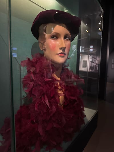 A mannequin is displayed in a glass case. The mannequin is positioned on its side, facing the camera. It has a pale complexion, with rouged cheeks and red lipstick. The mannequin wears a dark velvet beret and a red feather boa around its neck and shoulders. The background visible through the glass case appears to be a museum exhibition space with framed images on the walls. The lighting is dim, creating a moody atmosphere. The overall scene suggests a historical display or fashion exhibit.