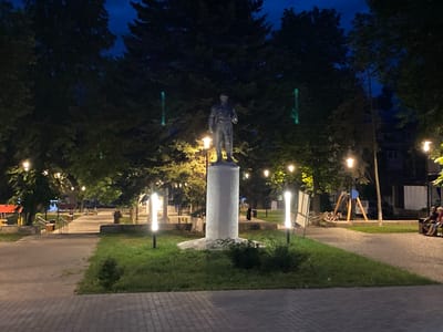 The image depicts a public park at dusk or night. In the foreground, a paved walkway leads into the park. To the right, a grassy area slopes down, and further back, another paved path is visible. Dominating the center of the frame is a tall, cylindrical pedestal, upon which stands a dark-colored statue of a man in a suit. The statue is illuminated by a lamp positioned at its base. Surrounding the pedestal is a lawn with bushes at its base. Several tall, modern-style lamps with vertical light sources are positioned along the pathways. In the background, mature trees with dense foliage create a dark canopy. Beyond the trees, the dimly lit facade of a building is visible, along with a playground area featuring a swing set and a slide. Several people are present in the park: a few individuals are seated on benches in the background to the right, and children are on the playground equipment. The sky is dark blue, indicating nighttime. Based on the presence of the statue of a man in a suit and the style of the park, this scene is likely located in the city of Antratsyt, Ukraine, potentially a memorial or historical site.