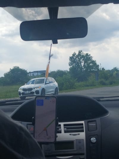 The image captures a view from inside a vehicle, looking forward. A white BMW SUV with license plate CSV-989 is visible on the road ahead. A smartphone displaying a navigation app is mounted on the dashboard in the foreground. The environment suggests an overcast day, with trees and buildings visible in the background. The location is identified as Budești, Moldova.
