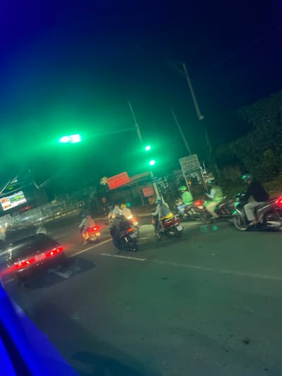 The image depicts a nighttime scene on a street in Binh Thuan Ward, Vietnam. A traffic light displays green, illuminating the area with a green hue. Several people are riding motorcycles, some wearing helmets. A black car is visible in the foreground, with its brake lights illuminated. In the background, there are buildings, trees, and a sign with Vietnamese text. The street is paved and appears to be an urban environment. The overall impression is one of traffic and movement at night.