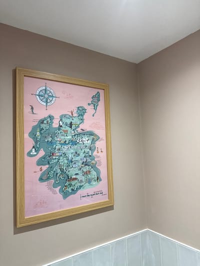 An upward-angled shot captures a large, framed, illustrated map hanging on a light taupe-colored wall in an indoor setting. The ceiling, painted a lighter off-white, is visible at the top, creating a corner where it meets the wall. To the right, another wall or structural element, featuring light grey horizontal tiles, is illuminated by a narrow strip of light, likely an LED.

The map, the central subject, is enclosed in a light-colored wooden frame with a visible grain. The map's background is a soft pink, with the landmasses depicted in a muted teal or green. It features a stylized, whimsical design with numerous small illustrations of landmarks, animals, and people. A prominent compass rose, in blues and white, with cardinal directions (N, E, S, W), is located in the bottom-left corner of the map.

Various detailed illustrations are visible across the map, including a person in a kilt, lighthouses, castles, traditional houses, a highland cow, deer, seals, and other flora and fauna. Swirling patterns suggestive of wind or water are also integrated into the design. A smaller, elongated island is depicted to the top-left of the main landmass. Towards the top-right, a scale bar indicates "100 KILOMETRES" and "100 MILES," with markings for 0, 50, and 100. While much of the smaller text denoting places or features is illegible due to distance, the overall artistic style suggests a map of a specific region, likely within Scotland, given visual cues like the kilt and highland cow, aligning with the Aberfoyle, United Kingdom context. No activity or event is taking place in this static scene.