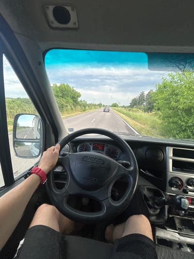 A first-person perspective photograph captures the view from inside an Iveco vehicle, likely a truck or large van, as it travels down a two-lane road in Byal Izvor, Bulgaria. The image is taken during bright daylight, showcasing clear skies with scattered clouds.

In the foreground, a person's right arm, wearing a distinctive red digital watch, holds the steering wheel, which prominently displays the "IVECO" logo. To the right, parts of the dashboard and a manual gear stick, showing a "R 1 3 5" and "2 4 6" pattern for a 6-speed transmission, are visible. The driver's legs, clad in black shorts, are also seen. The dashboard includes instrument clusters and a small digital display showing "2 Jun" and a number that looks like "053," possibly a trip meter.

Through the windshield, a paved road extends into the distance, lined on both sides by green trees and dense vegetation. A dark-colored vehicle can be seen far ahead on the road. The top part of the windshield has a noticeable blue tint. The right side mirror reflects the road stretching out behind the vehicle. The overall scene depicts a calm journey through a rural or semi-rural landscape.
