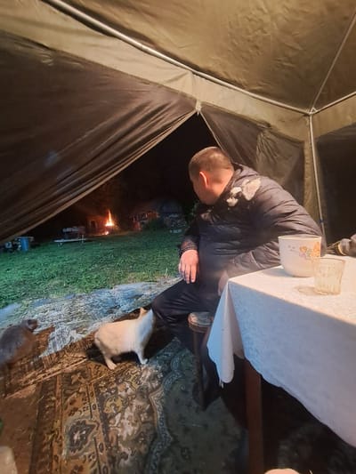 This media file captures an outdoor evening scene under a temporary shelter, likely a tent or gazebo.
**Subjects:**
The central figure is a man, seen from the back and side, seated at a white table. He has short hair and is wearing a dark, possibly puffy, jacket that appears to have some white residue on the left shoulder. His left arm is visible, resting near the table. On the white tablecloth, there is a white ceramic mug with a floral/fruit design and a clear glass. The structure he is in is a large, light-brown or olive-colored tent, with visible metal poles forming its frame. On the ground beneath the table and extending outwards, there is a patterned rug with intricate designs in shades of brown, beige, and darker tones. Two animals are visible near the table; one light-colored, resembling a cat or small dog, and another darker, less distinct animal in the lower-left foreground. In the background, beyond the tent structure, an area of green grass is visible, and a small bonfire or open flame burns in the distance.
**Activity or event taking place:**
The man appears to be resting or having a meal/drink at the table within the shelter. The bonfire in the background suggests a casual outdoor gathering or a leisurely evening activity.
**Notable details:**
The scene is set at night or during very low light conditions, indicated by the darkness outside the illuminated immediate area and the prominent glow of the distant bonfire. The man's puffy jacket suggests cool temperatures. Parts of the ground, including sections of the rug and grass, appear damp or wet. There is no clearly visible text in the image.
**Location context:**
The event takes place in Polianki, Russia.