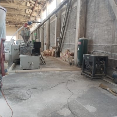 The image shows the interior of an industrial facility, likely a factory or processing plant, located in Iskandar, Uzbekistan. The environment is characterized by concrete floors and brick walls, with exposed structural beams and a high ceiling. Various pieces of industrial machinery are visible, including what appears to be a large grinder or mixer, a generator set, and an air compressor tank. Bags of material are stacked along the walls, suggesting ongoing production or storage. Light enters the space from windows in the upper part of the building. Several cables and pipes are present on the floor and along the walls, indicating the presence of active or recently used equipment. The scene appears somewhat dusty, with scattered material on the floor, and the overall impression is one of a functional but perhaps older industrial setting. There are no people visible in the image.