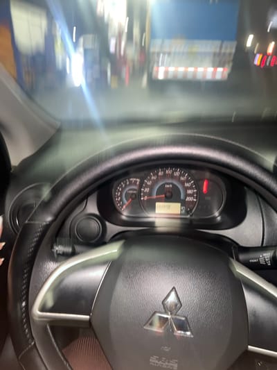 The image shows the interior of a Mitsubishi car, focusing on the steering wheel and instrument panel. The dashboard displays a speedometer reading indicating the car is not in motion, with an odometer reading of 062608. The background appears blurry, suggesting the photo was taken while in motion. The surroundings are indistinct, making it difficult to pinpoint a specific location. However, given the provided information, it is supposed to be in the city of Lurín, Peru.