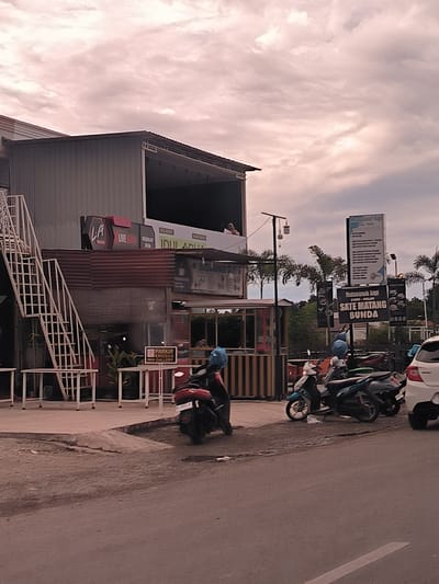 The image presents an outdoor street scene in Keuniree, Indonesia, under an overcast sky, likely in the late afternoon or early evening due to the soft, warm light. The central feature is a two-story commercial building with an industrial aesthetic, characterized by corrugated metal walls. An external white metal staircase with railings provides access to the upper level, where a person is subtly visible on an open-air balcony.

On the ground floor, an outdoor seating area with white tables and chairs is set up. Several motorcycles of various colors, including red, blue, and black, are parked on the paved area in front of the building. A white car is partially visible on the far right. Palm trees dot the background, hinting at a tropical climate.

Various signs advertise businesses. Prominently displayed text includes "LA," "LIVE," and "BERKAT DUA" on signs attached to the building. Another sign directs attention to "SATE MATANG BUNDA," likely a food establishment, with "MALEM" partially visible. A smaller sign clearly indicates "PARKIR KHUSUS MMA GALLERY" (Special Parking MMA Gallery). Other visible text snippets include "SELECT" and "IDUL ADHA." The scene conveys a quiet, daily street ambiance with commercial activity.