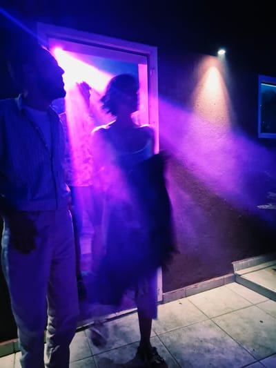 This is a low-light, motion-blurred photograph taken at night in Herceg Novi, Montenegro. The scene appears to be an outdoor area near an entrance, possibly a doorway or a patio. Two individuals are visible, both heavily blurred due to movement and low light conditions.
In the foreground left, a person is seen in profile, facing towards the right of the frame. This individual is wearing a light-colored, vertically striped long-sleeved shirt and light-colored trousers. Their facial features are indistinct due to motion blur, but a beard is discernible. They appear to be standing still or moving slowly.
To the right of this person and slightly behind them, another individual is in motion, creating significant motion blur. This person is wearing a dress or a similar garment that appears to be light in color. They are holding an object, possibly a dark bag or jacket, in their right arm.
The environment is characterized by strong purple and pink lighting, likely from stage lights or decorative lighting, creating dramatic shafts of light that illuminate the smoke or haze in the air. A rough, textured wall with a light fixture casting a yellow-orange beam is visible on the right side of the frame. The ground is tiled, and a set of steps is visible in the background to the right. The overall impression is that of a lively, possibly festive, outdoor gathering or event. The location within Herceg Novi is not explicitly identifiable from the visual cues alone, but the atmospheric lighting and the setting suggest a public or semi-public space.