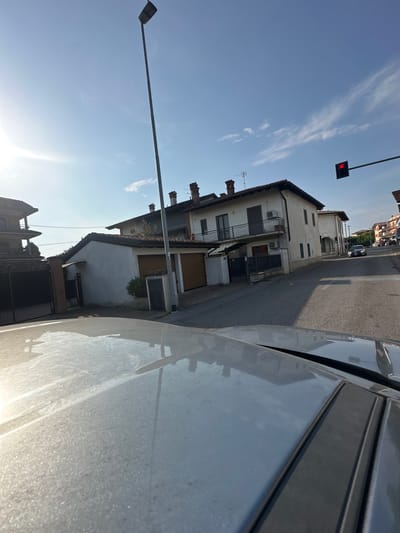The image depicts a street scene in Rudiano, Italy. The perspective is from inside a vehicle, looking forward and slightly upwards. The foreground shows the hood of a light-colored car, dusted with a fine layer of particles, reflecting the sky and surrounding buildings.
In the mid-ground, a paved road curves to the right, with houses and a garage visible on the left and right sides. The houses appear to be two-story residential buildings with tiled roofs and balconies. One house on the left has a dark metal gate and a brick pillar. Another house to the right, painted white, features multiple windows and balconies. Adjacent to this house is a garage with a light brown, segmented door. Further down the street, other buildings and trees can be seen.
In the background, the sky is a clear blue with some wispy white clouds and a bright sun is visible on the left side. A tall, dark lamppost stands prominently on the left side of the street. A traffic light with a red illuminated signal is positioned above the road on the right side, attached to a horizontal arm extending from a pole. A dark-colored car is driving on the road in the distance, approaching the intersection controlled by the traffic light. The overall impression is a sunny day in a residential area.