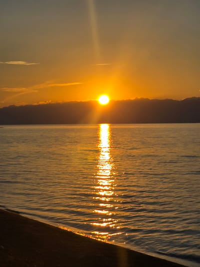 This is a photograph of a sunset over a body of water. The sun is a bright, orange orb, low on the horizon, casting a golden reflection across the rippling surface of the water. The sky is a hazy orange and yellow, with some wispy clouds. On the right side of the image, a dark, angled object juts into the frame, creating a strong contrast with the luminous water and sky. The overall mood is tranquil and serene, evoking the peacefulness of a sunset. The scene appears to be a natural landscape, likely a lake or the sea. The time of day is clearly sunset, indicated by the position and color of the sun and the warm lighting. There is no visible text in the image.