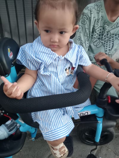 A toddler sits on a light blue tricycle in Bungur, Indonesia. The child wears a blue and white striped shirt with a Mickey Mouse design, and tan sandals. The tricycle has a black padded safety bar and a black handlebar. An older child or adult stands behind, holding the handlebar and wearing a light green shirt with a black and white polka dot design. The setting appears to be outdoors, possibly on a paved surface with a fence in the background.