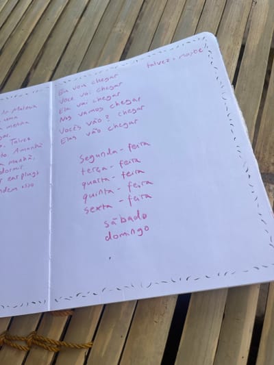 An open white-paged notebook rests on a light brown, slatted bamboo surface, viewed from an overhead perspective, in Desa Batu Layar, Indonesia. The notebook contains handwritten text in magenta ink across both visible pages. The right page displays Portuguese language learning notes, including verb conjugations such as "Eu vou chegar" (I will arrive) and a list of the days of the week from "Segunda-feira" (Monday) to "Domingo" (Sunday), with "talvez = maybe" noted at the top right. A hand-drawn dashed line forms a decorative border along the edges of both pages. Below the notebook, a section of yellow braided rope, secured with visible knots, is present on the bamboo surface. No individuals or active interactions are depicted within the scene.