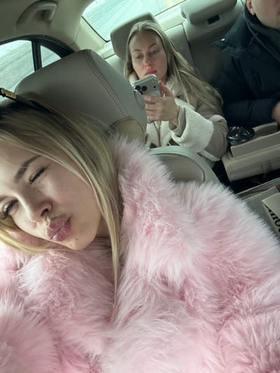 The image captures three individuals inside the rear cabin of a passenger vehicle, reportedly located in the city of Bratovshchina, Russia. In the immediate foreground, a young blonde female, partially visible, is taking a selfie. She has light skin, visible freckles, and long blonde hair, with her lips pursed in a "kissing" expression and eyes partially closed. She is wearing a prominent, light pink fluffy garment that dominates the foreground. Directly behind her, a second blonde female is seated, facing forward, engrossed with a light-colored smartphone held in both hands. Her long, straight blonde hair is visible, and she is wearing a light-beige or tan coat featuring a white, fluffy lining on the cuffs. To the right of this second female, a third individual, identified as male, is partially visible from the shoulder up, dressed in a dark jacket. The vehicle's interior consists of light-colored seats and headrests. A blurry, indistinct outdoor environment, possibly an overcast sky, is visible through the rear passenger window on the left. A small, dark circular object rests on a console or armrest between the two rear passengers.