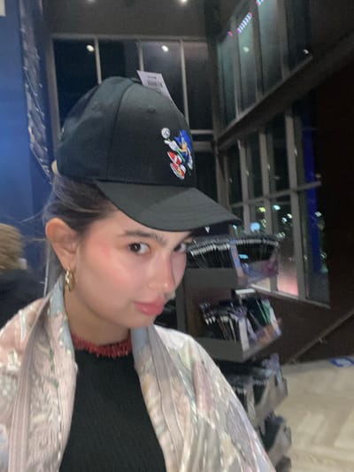 A woman is pictured wearing a black baseball cap with a Sonic the Hedgehog character embroidered on the side. She has a fair complexion, light brown hair peeking out from under the cap, and is wearing earrings and a red beaded necklace. The top she's wearing appears to be black, and she has a light-colored patterned scarf or wrap around her shoulders. The background shows clothing racks and displays suggesting a retail environment, possibly a store. The photo appears to have been taken indoors.