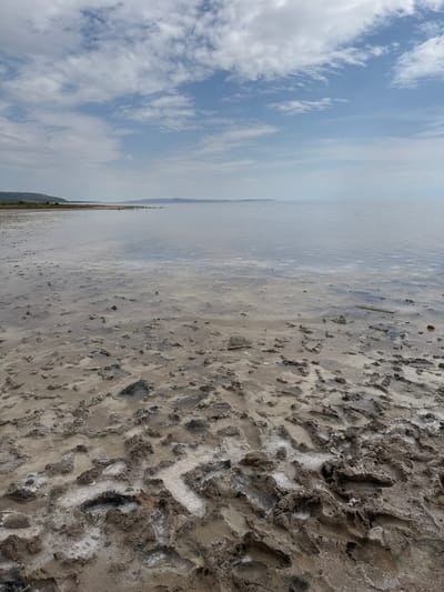 The image depicts a landscape with a sky filled with clouds on the left side and a vast body of water on the right. The ground on the right is a muddy or sandy surface with numerous footprints, suggesting recent human activity. This is the dried out beach of Lake Tuz, which is the second largest lake in Turkey. The city in the background is Şereflikoçhisar in Ankara province. The overall impression is of a natural environment with evidence of human presence.