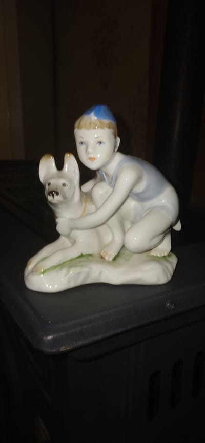 A porcelain figurine of a young boy in a blue cap and a light blue outfit is posed with a white dog. The boy is kneeling and embracing the dog, which is lying down. The figurine is placed on a dark, textured surface, possibly the top of a piece of furniture or an appliance, against a dark background. The overall lighting is dim. There is no discernible connection to Ganja City, Azerbaijan.
