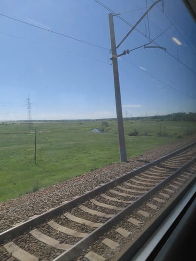 The image shows a view from a train window, looking out at a rural landscape. The foreground features a set of train tracks, with ballast and concrete sleepers visible. To the left of the tracks is a grassy field, dotted with a few small trees and a single wooden pole. In the middle ground, the field continues, with a narrow body of water and several power pylons visible in the distance. The background is dominated by a clear blue sky. The location is identified as Chik, Russia. There are no people or animals visible in the image.